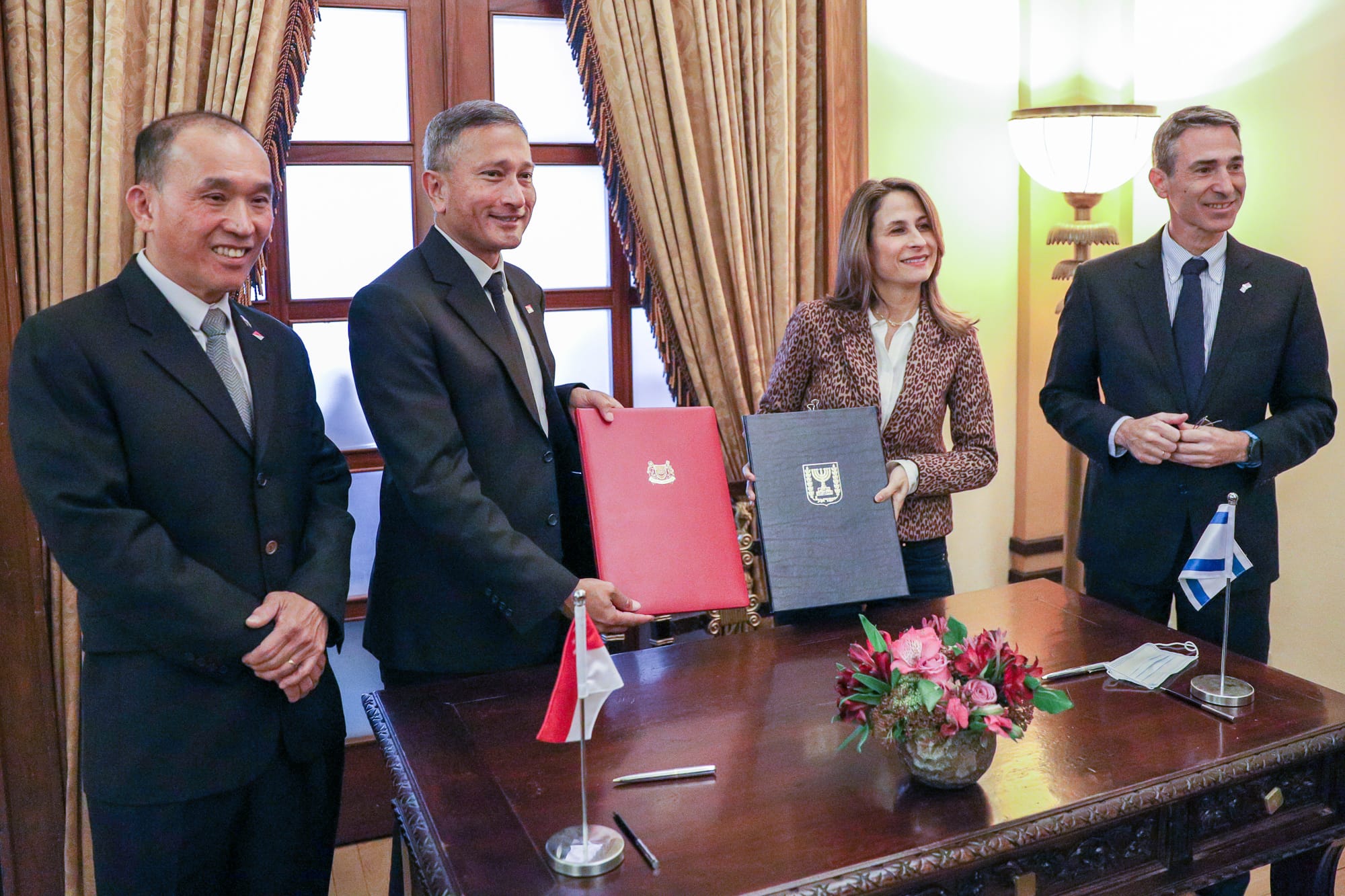 Four people in suits holding documents; Singapore and Israeli flags on table.